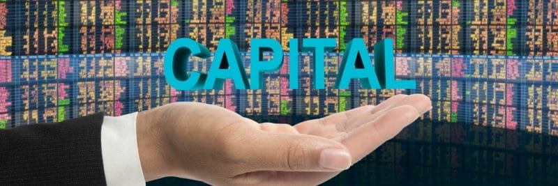 picture of a hand holding the word capital with financial screens in the background
